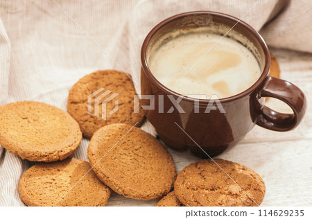 A cup of aromatic coffee cappuccino with Oatmeal cookies. Morning rays and latte for breakfast in a beautiful brown cup. Wooden background. A cup of aromatic coffee cappuccino with Oatmeal cookies. Morning rays and latte for breakfast in a beautiful brown cup. Wooden background. 114629235