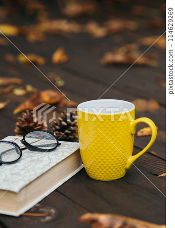 Yellow cup of tea with a book and glasses on a wooden background with a lot of dry fallen leaves. Relaxing scene. Selective focus. Bad weather 114629269