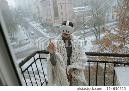 On the balcony in winter, a girl embraces virtual reality adventures with a headset. 114630813