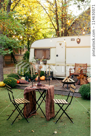 Autumn evening, romantic dinner in the garden. Table with tablecloth and decoration. Trailer with autumn decor and light bulbs 114630871