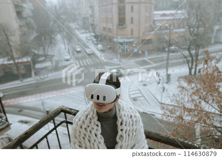 Immersed in virtual reality, a girl wears a VR headset on the balcony during winter. 114630879