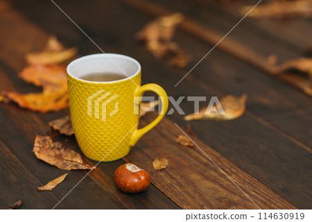 Yellow cup of tea and chestnut at wooden background with a lot of red and yellow fallen leaves. Relax scene. Selective focus and copy space. 114630919