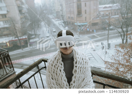 Enjoying VR experiences, a young lady dons a headset on the winter balcony. 114630920