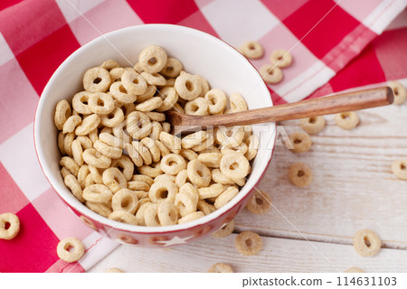 healthy cereal rings on a light wood background with tablecloth red in a plate and scattered on the table with sun rays. morning eat. Healthy children's breakfast. Dry muesli. healthy cereal rings on a light wood background with tablecloth red in a plate and scattered on the table with sun rays. morning eat. Healthy children's breakfast. Dry muesli. 114631103