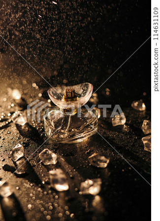 A bottle of women's perfume in the rays of golden light on a black background with ice cubes, water drops and splashes. Play of light on a bottle of perfume A bottle of women's perfume in the rays of golden light on a black background with ice cubes, water drops and splashes. Play of light on a bottle of perfume 114631109