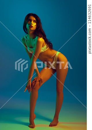 Full-length portrait of sexy young girl with brunette hair and plump lips, slim body posing in tender lingerie against cyan background in neon light Full-length portrait of sexy young girl with brunette hair and plump lips, slim body posing in tender lingerie against cyan background in neon light 114632691
