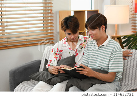 Relaxed young gay couple reading book on couch at home. LGBTQ, leisure and lifestyle concept Relaxed young gay couple reading book on couch at home. LGBTQ, leisure and lifestyle concept 114632721