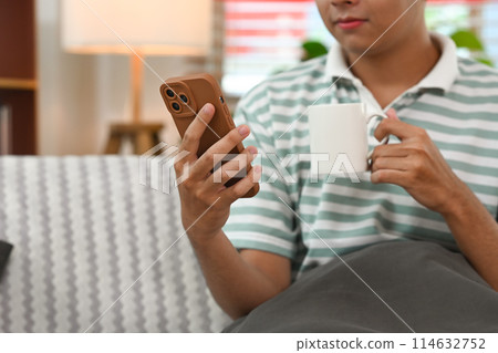Young smiling man holding smartphone and drinking coffee relaxing on couch at home Young smiling man holding smartphone and drinking coffee relaxing on couch at home 114632752