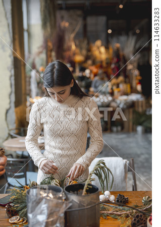 A young woman crafting a Christmas wreath at a holiday decor workshop. 114633283