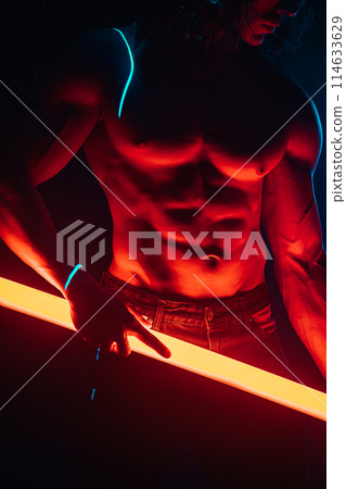 Muscular strong guy with naked torso abs holds neon light tube in dark room Muscular strong guy with naked torso abs holds neon light tube in dark room 114633629