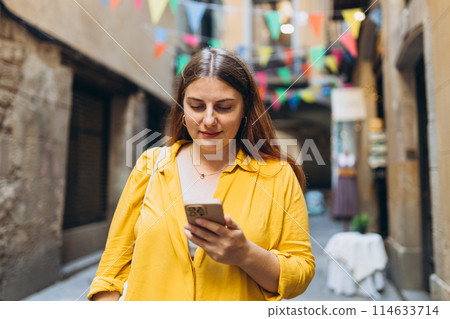 30s woman tourist dressed in casual look holding smartphone gadget in hand for communicate on the city street. Happy woman chatting on phone and relaxing outdoors. Urban, people concept. Old town 114633714