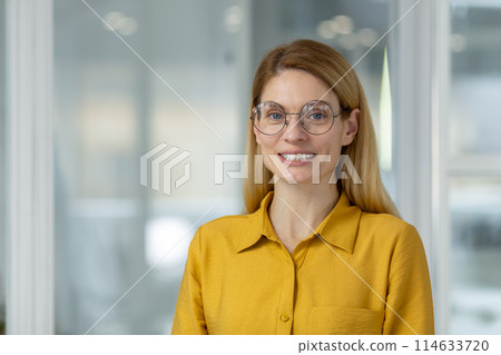 Confident and happy female professional in a yellow shirt and glasses smiling in a modern office environment. Confident and happy female professional in a yellow shirt and glasses smiling in a modern office environment. 114633720