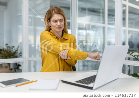 A woman in a yellow shirt experiencing elbow pain while working on her laptop in a modern office environment. A woman in a yellow shirt experiencing elbow pain while working on her laptop in a modern office environment. 114633737