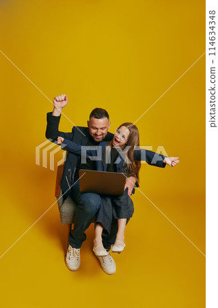 Business Partners. dad and his daughter collaborate over laptop, with joyful expression raising hands against sunny-yellow background. Business Partners. dad and his daughter collaborate over laptop, with joyful expression raising hands against sunny-yellow background. 114634348