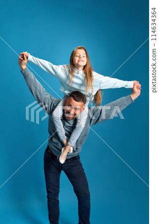Fly together. Little child, girl sitting on father's shoulders and playing with him airplanes against blue studio background. 114634364