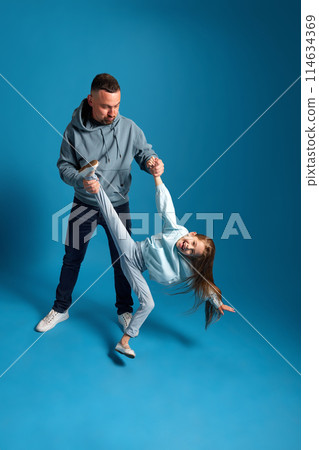 Father playing with his little daughter, holding her by hand and leg in air against blue studio background. 114634369