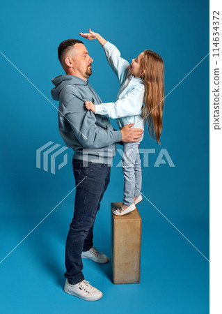 Portrait of little girl stands on stand and measures her height with her father, showing how she grew up against blue background. 114634372