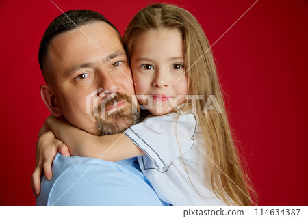 Portrait of young man holds on hands his beloved little girl against vibrant red studio background. Dad's daughter. 114634387