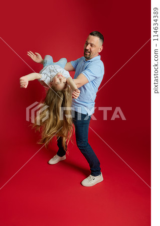 Father-Daughter Playtime. Man joyfully lifts his daughter high, her smile wide with excitement against vibrant red background. 114634389