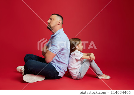 Father and daughter sits back-to-back with thoughtful expression against vibrant red background wit negative space. 114634405