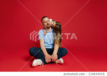 Father smiling while his beloved little daughter kissing and hugging his behind back against vibrant red background. 114634419