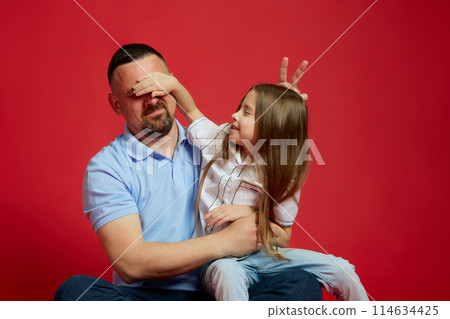 Playful moments. Charming little girl covered her father eyes while they posing against vibrant red studio background. Playful moments. Charming little girl covered her father eyes while they posing against vibrant red studio background. 114634425