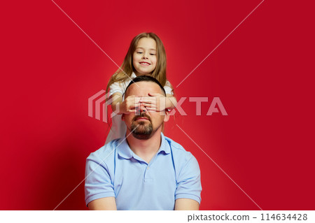 Heartwarming moments. Father sits and his little daughter covered his eyes from behind against vibrant red background. 114634428