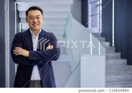 Confident asian mature man in business attire standing with arms crossed in modern office building background, showcasing success and professionalism. 114634884