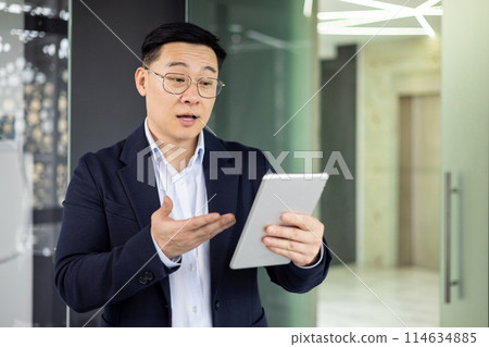 Asian mature man in business attire using a tablet, discussing or presenting in a modern office setting, showcasing technology and business communication. 114634885