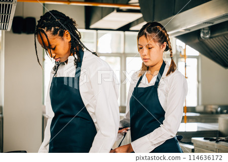 Girlfriend chef helps woman friend tie apron in a restaurant kitchen. Two adults in uniform prepare for food education standing with a painting ready for service. Girlfriend chef helps woman friend tie apron in a restaurant kitchen. Two adults in uniform prepare for food education standing with a painting ready for service. 114636172
