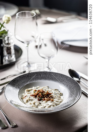 Halusky as traditional Slovak potato gnocchi with sheep cheese bryndza, fried bacon and chives served in a modern plate and on a festively laid restaurant table. 114636720