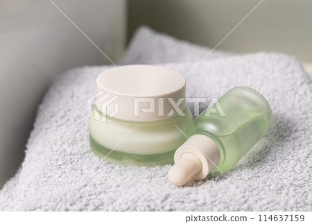 Light green dropper bottle and cream jar on light grey bath towel near basin closeup 114637159