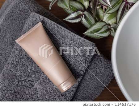 Beige cosmetic tube on grey folded towel near green plant on wood in bathroom, mockup Beige cosmetic tube on grey folded towel near green plant on wood in bathroom, mockup 114637176