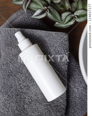 White spray cosmetic bottle on grey folded towel near green plant on wood in bathroom, mockup 114637177