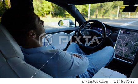Male businessperson experience to ride an autonomous self driving electric car at urban road. Confident businessman resting during riding on electrical auto with autopilot at city. Slow motion Male businessperson experience to ride an autonomous self driving electric car at urban road. Confident businessman resting during riding on electrical auto with autopilot at city. Slow motion 114637588
