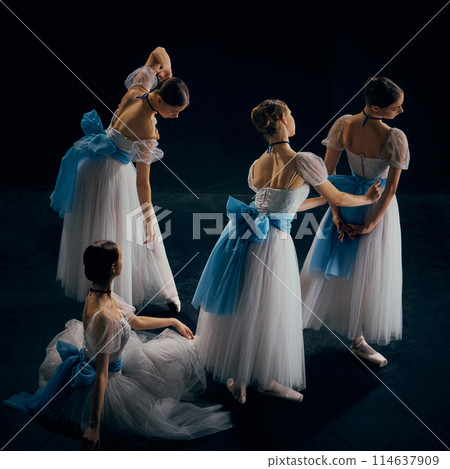 Group of ballerinas dressed in elegant white tutus with delicate blue sashes, practicing their graceful poses on dimly lit stage 114637909