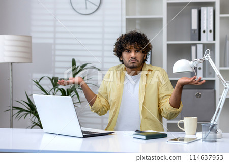 Young man looking confused, shrugging shoulders with uncertainty while working on a laptop in an office environment. 114637953