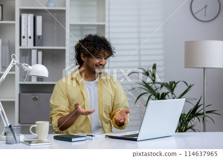 Smiling man in a yellow shirt engaging in a video call while sitting at a desk with a laptop in a bright, well-organized office. 114637965
