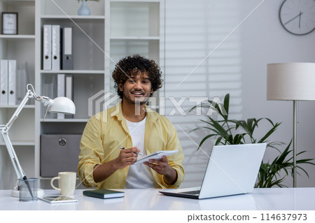 A cheerful man participates in an online course, taking notes while watching a webinar on a laptop in a modern home office. A cheerful man participates in an online course, taking notes while watching a webinar on a laptop in a modern home office. 114637973