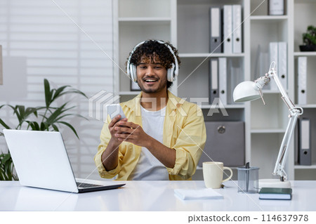 Smiling man using a smartphone and listening to music with headphones while sitting at his home office desk with laptop and coffee. 114637978