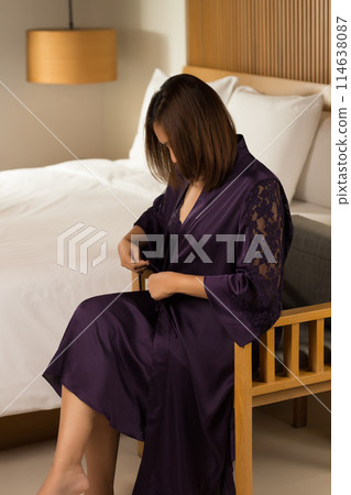 Asian lady is perched on a wooden chair, adorning herself with a luxurious silk gown in her private quarters. 114638087