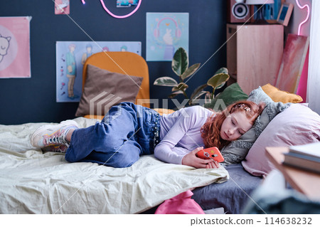 Modern Caucasian teen girl spending day indoors relaxing on bed in her bedroom watching videos in social media on smartphone 114638232
