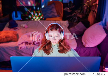 High angle view of modern teen girl wearing headphones sitting in her room surfing Internet on desktop computer and listening to music 114638317