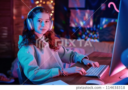 Portrait of confident Caucasian girl wearing hoodie and headphones sitting at desk in her room looking at camera while playing video game on computer 114638318