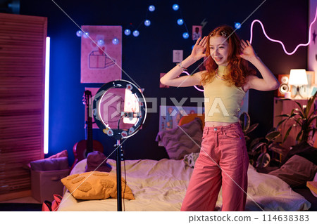 Modern teen influencer standing in neon bedroom posing for smartphone camera while recording video for blog, copy space Modern teen influencer standing in neon bedroom posing for smartphone camera while recording video for blog, copy space 114638383