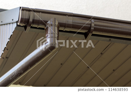 Close-up detail of cottage house corner with brown metal planks siding and roof with steel gutter rain system. Roofing, construction, drainage pipes installation and connection concept. Close-up detail of cottage house corner with brown metal planks siding and roof with steel gutter rain system. Roofing, construction, drainage pipes installation and connection concept. 114638593