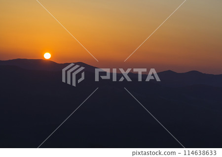 Beautiful sunset in mountains. Wide panorama view of big bright white sun in dramatic orange sky over dark mountain range landscape at sunset or sunrise. Beauty of nature concept. 114638633