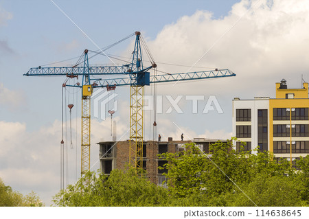 Urban view of silhouettes of two high industrial tower cranes working at construction of new brick building with workers in hard hats on it against bright blue sky and green top trees background. 114638645