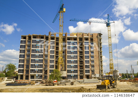 Building site with two industrial tower cranes working at construction of new brick tall building with a yellow tractor bulldozer on bright blue sky and green trees background. Building site with two industrial tower cranes working at construction of new brick tall building with a yellow tractor bulldozer on bright blue sky and green trees background. 114638647