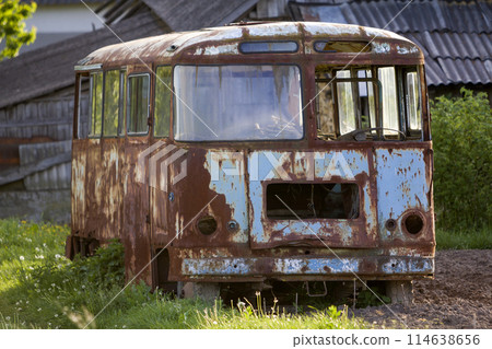 Close-up of old forsaken passenger bus with broken windows rusting in high green weedy grass on edge of plowed brown field on bright spring day under blue morning sky. Close-up of old forsaken passenger bus with broken windows rusting in high green weedy grass on edge of plowed brown field on bright spring day under blue morning sky. 114638656